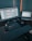 A workspace with a large monitor displaying a design program and a laptop showing a website. On the desk, there is a wireless keyboard, a mouse on a mouse pad, a smartphone, a red pencil, and a small bottle of eye drops. Soft lighting casts shadows on the wall behind the setup.
