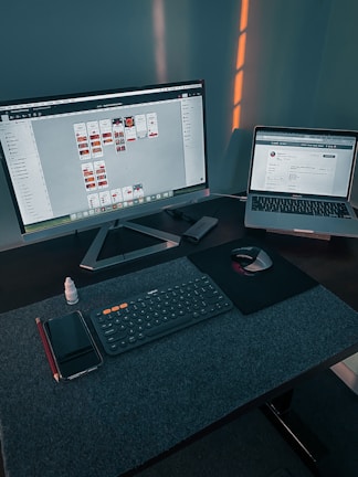 A workspace with a large monitor displaying a design program and a laptop showing a website. On the desk, there is a wireless keyboard, a mouse on a mouse pad, a smartphone, a red pencil, and a small bottle of eye drops. Soft lighting casts shadows on the wall behind the setup.