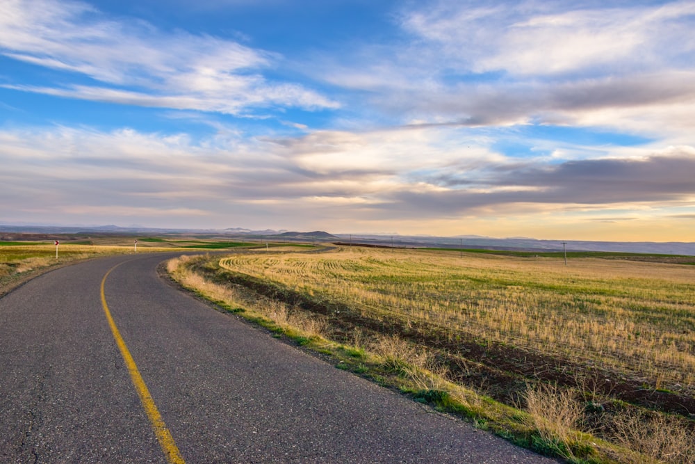 gray concrete road between green grass field under white clouds and blue sky during daytime