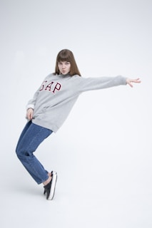 A person in a casual, dynamic pose wearing a grey sweatshirt with the word 'GAP' printed in red letters. They also wear blue jeans and black sneakers, standing against a plain white background.