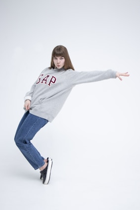 A person in a casual, dynamic pose wearing a grey sweatshirt with the word 'GAP' printed in red letters. They also wear blue jeans and black sneakers, standing against a plain white background.
