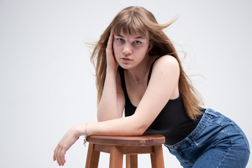 A young person with long brown hair wearing a black sleeveless top and blue jeans leans against a wooden stool. The expression on their face appears contemplative or introspective, and the background is plain white.