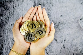 A pair of hands holding a collection of golden coins with the Bitcoin symbol. The background is a textured gray surface.