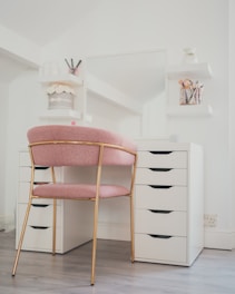 A well-organized vanity area featuring a white desk with multiple drawers and a large mirror. A pink cushioned chair with gold metal legs is positioned in front of the desk. The area is decorated with cosmetic brushes in a holder, jars, and small vases on two white floating shelves attached to the wall.