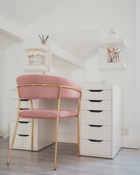 A well-organized vanity area featuring a white desk with multiple drawers and a large mirror. A pink cushioned chair with gold metal legs is positioned in front of the desk. The area is decorated with cosmetic brushes in a holder, jars, and small vases on two white floating shelves attached to the wall.