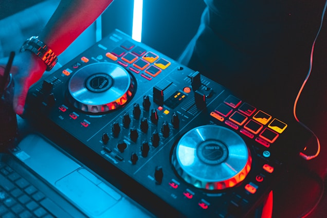 A close-up view of a DJ controller with various buttons, knobs, and two large jog wheels. The setup is illuminated with vibrant red and blue LED lights. A person's hand is visible, suggesting interaction with the equipment, and a laptop is partially visible on the side, indicating a digital music setup.
