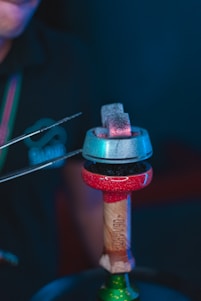 A close-up of a hookah setup, featuring red-hot charcoal pieces glowing atop a metallic heat management device. The setup is placed on a colorful, textured hookah bowl, which sits on the stem. Tongs are visible, holding one of the charcoal pieces.
