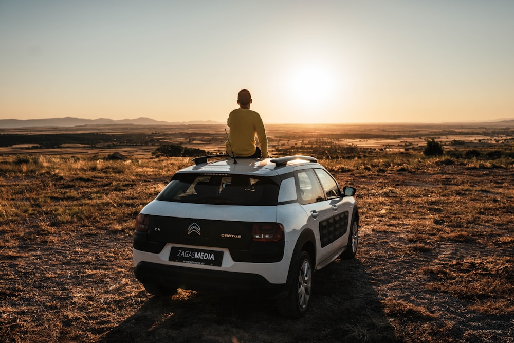 man in brown jacket sitting on white jeep wrangler on brown field during daytime