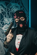 A person wearing a black balaclava with pink embroidery and holding a handgun. The person is dressed in a black outfit with tattoos visible on their hand and wrist. Behind them is a geometric, abstract wall design featuring a face pattern.