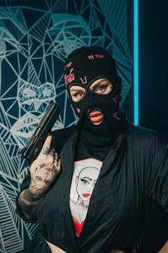 A person wearing a black balaclava with pink embroidery and holding a handgun. The person is dressed in a black outfit with tattoos visible on their hand and wrist. Behind them is a geometric, abstract wall design featuring a face pattern.