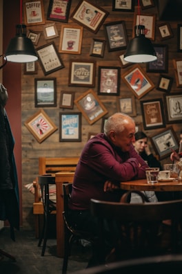 A cozy cafe setting with wooden walls adorned with numerous framed coffee-themed pictures and posters. Two individuals are seated at a table, with one person prominently in view, appearing thoughtful. The furnishings include wooden chairs and tables, and overhead, there are black pendant lamps providing warm lighting.