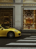A luxury storefront with gold accents featuring a window display of designer items. A vibrant yellow sports car is parked in front, and a zebra crossing is visible on the street.