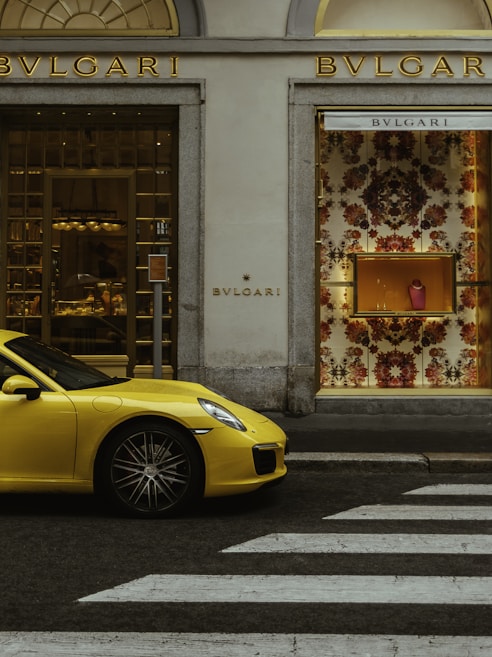 A luxury storefront with gold accents featuring a window display of designer items. A vibrant yellow sports car is parked in front, and a zebra crossing is visible on the street.