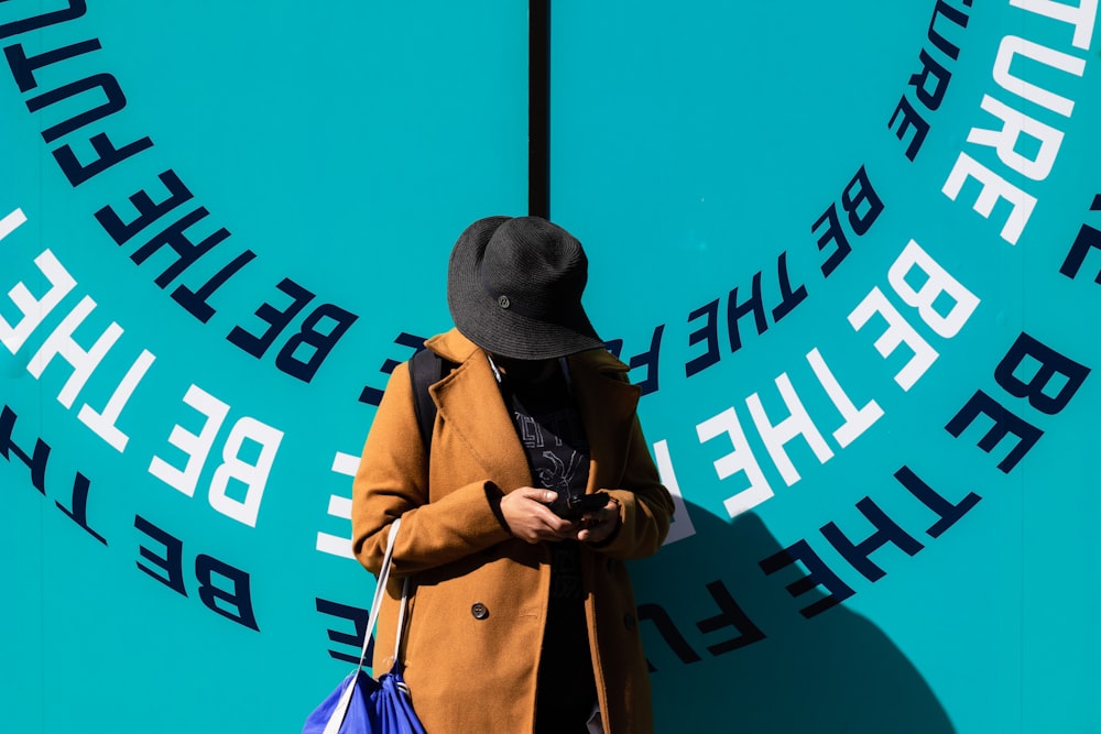 man in brown coat wearing black fedora hat standing beside blue and white wall