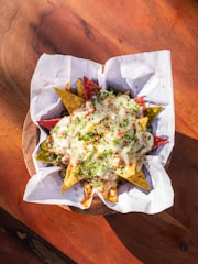 A basket filled with colorful tortilla chips covered in melted cheese and garnished with herbs. The chips are served on a wooden surface with a lining of white parchment paper.