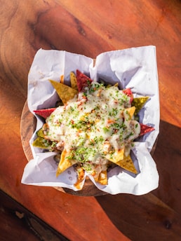 A basket filled with colorful tortilla chips covered in melted cheese and garnished with herbs. The chips are served on a wooden surface with a lining of white parchment paper.