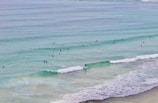Numerous surfers are scattered across a calm ocean, waiting to catch waves. The water is a clear mix of turquoise and blue hues, and gentle waves roll towards the sandy shore. The overall view suggests a relaxed atmosphere perfect for a day of surfing.