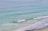 Numerous surfers are scattered across a calm ocean, waiting to catch waves. The water is a clear mix of turquoise and blue hues, and gentle waves roll towards the sandy shore. The overall view suggests a relaxed atmosphere perfect for a day of surfing.