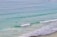 Numerous surfers are scattered across a calm ocean, waiting to catch waves. The water is a clear mix of turquoise and blue hues, and gentle waves roll towards the sandy shore. The overall view suggests a relaxed atmosphere perfect for a day of surfing.