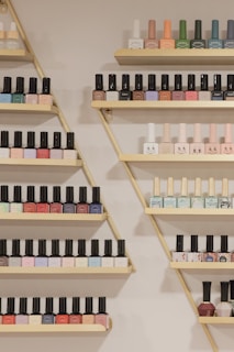 A neatly arranged display of nail polish bottles on triangular wooden shelves, featuring a variety of colors such as pastels, bright shades, and darker tones. The bottles are organized by color and brand, creating a visually appealing pattern.