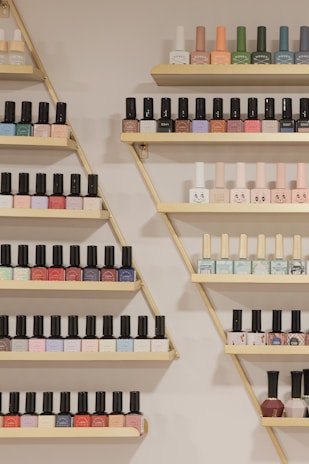A neatly arranged display of nail polish bottles on triangular wooden shelves, featuring a variety of colors such as pastels, bright shades, and darker tones. The bottles are organized by color and brand, creating a visually appealing pattern.