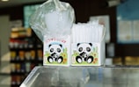 Two boxes featuring a cartoon panda design are placed on a glass counter. One box contains white plastic cups wrapped in a transparent plastic, and the other holds clear, plastic straws. The background is a slightly blurred interior, suggesting a convenience store or similar setting with shelves displaying colorful products.
