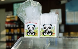 Two boxes featuring a cartoon panda design are placed on a glass counter. One box contains white plastic cups wrapped in a transparent plastic, and the other holds clear, plastic straws. The background is a slightly blurred interior, suggesting a convenience store or similar setting with shelves displaying colorful products.
