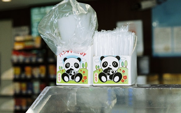 Two boxes featuring a cartoon panda design are placed on a glass counter. One box contains white plastic cups wrapped in a transparent plastic, and the other holds clear, plastic straws. The background is a slightly blurred interior, suggesting a convenience store or similar setting with shelves displaying colorful products.