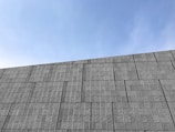 A concrete wall with a pattern of rectangular panels rises against a clear blue sky. The wall's surface has a textured appearance, providing a sense of depth and detail.