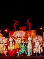 A group of performers in elaborate, traditional costumes stand on stage. The scene is rich with ornate headdresses, detailed costumes in vibrant colors, and intricate accessories. The backdrop features silhouettes and decorative elements, enhancing the theatrical setting. Bright lighting highlights the colorful costumes, suggesting a festive or cultural event.