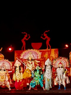 A group of performers in elaborate, traditional costumes stand on stage. The scene is rich with ornate headdresses, detailed costumes in vibrant colors, and intricate accessories. The backdrop features silhouettes and decorative elements, enhancing the theatrical setting. Bright lighting highlights the colorful costumes, suggesting a festive or cultural event.