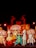 A group of performers in elaborate, traditional costumes stand on stage. The scene is rich with ornate headdresses, detailed costumes in vibrant colors, and intricate accessories. The backdrop features silhouettes and decorative elements, enhancing the theatrical setting. Bright lighting highlights the colorful costumes, suggesting a festive or cultural event.