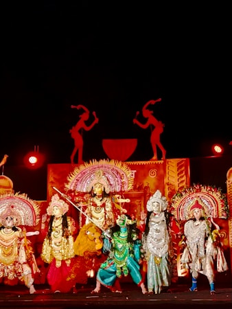 A group of performers in elaborate, traditional costumes stand on stage. The scene is rich with ornate headdresses, detailed costumes in vibrant colors, and intricate accessories. The backdrop features silhouettes and decorative elements, enhancing the theatrical setting. Bright lighting highlights the colorful costumes, suggesting a festive or cultural event.