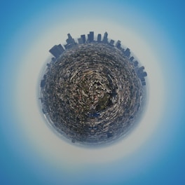 A panoramic view of a city presented in a circular, spherical format, giving the impression that the cityscape is encapsulated on a small planet. Tall skyscrapers are prominently visible around the edge, while smaller buildings and streets fill the interior.