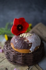 A round loaf of glazed bread placed on a wicker tray with a bright red flower on top. The bread has a mix of dark and light brown, topped with icing and some small nuts or seeds. In the background, there are blurred flowers and a textured, dark green background, along with some scattered old newspaper pages.