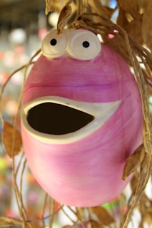 A whimsical ceramic sculpture with a round, pink body and a wide open mouth resembling a surprised expression. The eyes are large and cartoonish, positioned on top of the head, contributing to the playful appearance. Dried twigs or vines are attached to the top, adding an organic touch to the otherwise smooth surface of the sculpture.