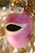 A whimsical ceramic sculpture with a round, pink body and a wide open mouth resembling a surprised expression. The eyes are large and cartoonish, positioned on top of the head, contributing to the playful appearance. Dried twigs or vines are attached to the top, adding an organic touch to the otherwise smooth surface of the sculpture.