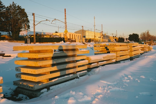 Stacked concrete beams are lined up in a snowy, industrial setting with train tracks visible in the background. The sun casts long shadows, highlighting the golden tones of the beams against the white snow. Buildings and trees are situated in the distance.