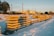 Stacked concrete beams are lined up in a snowy, industrial setting with train tracks visible in the background. The sun casts long shadows, highlighting the golden tones of the beams against the white snow. Buildings and trees are situated in the distance.