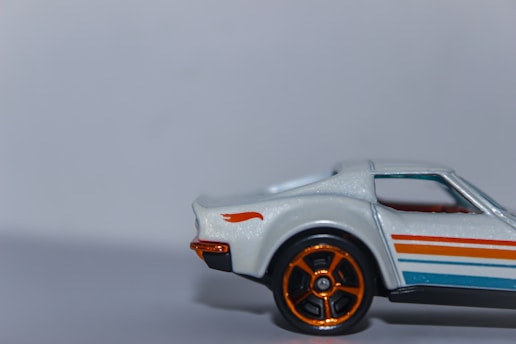 A close-up of a toy car model, featuring a sleek design with a white body and colorful stripes. The car has orange accents on the wheels and small details, with the rear section prominently visible against a plain background.