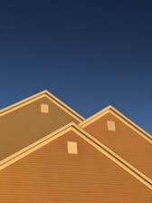 A series of house rooftops illuminated by golden sunlight against a backdrop of clear deep blue sky. The triangular roof peaks are prominent, showcasing the distinct lines and ventilation panels.