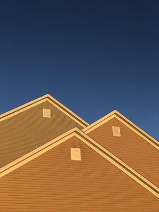A series of house rooftops illuminated by golden sunlight against a backdrop of clear deep blue sky. The triangular roof peaks are prominent, showcasing the distinct lines and ventilation panels.