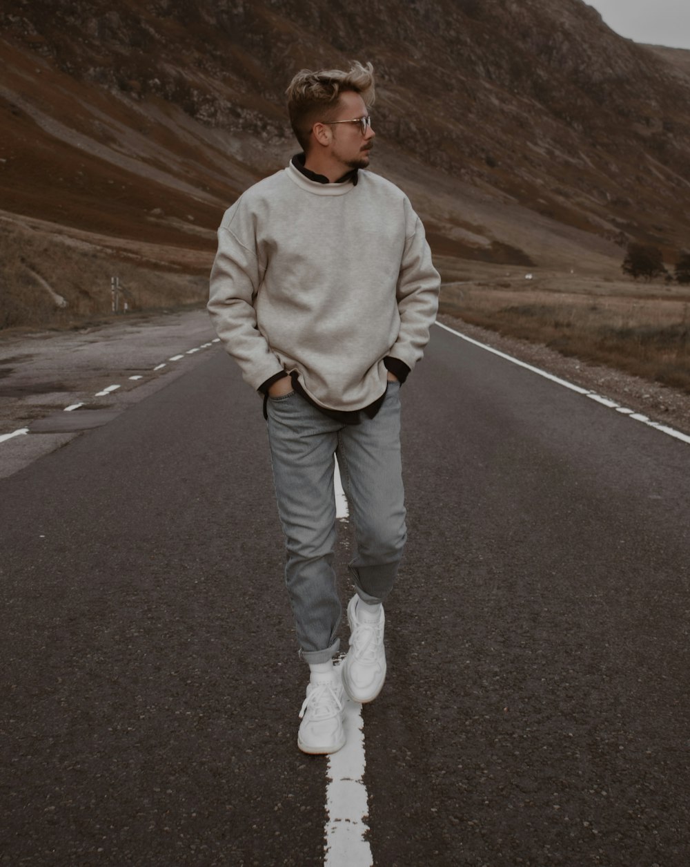 man in white sweater and blue denim jeans standing on gray asphalt road during daytime