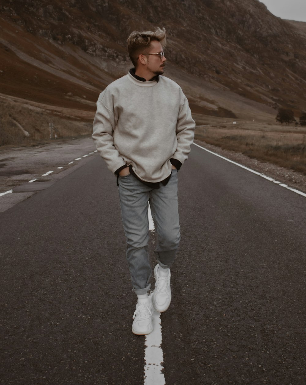 uomo in maglione bianco e jeans blu denim in piedi sulla strada asfaltata grigia durante il giorno