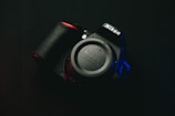 A Nikon camera rests against a dark background, with the brand name prominently visible on both the body and lens cap. Soft red and blue lighting highlights parts of the camera, creating a dramatic and moody effect.