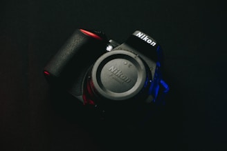 A Nikon camera rests against a dark background, with the brand name prominently visible on both the body and lens cap. Soft red and blue lighting highlights parts of the camera, creating a dramatic and moody effect.