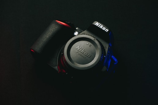 A Nikon camera rests against a dark background, with the brand name prominently visible on both the body and lens cap. Soft red and blue lighting highlights parts of the camera, creating a dramatic and moody effect.