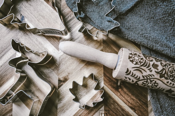 Several metal cookie cutters in shapes like a reindeer and maple leaf are arranged on a wooden surface beside a decorated wooden rolling pin. A textured dark cloth lies nearby, adding a rustic charm to the scene.