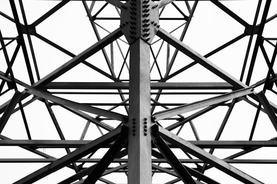 The image features the intricate metal framework of a structure, with various intersecting beams and bolts visible. The geometric patterns created by the beams form a complex lattice design.