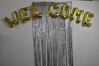 Gold foil balloons spell out the word 'WELCOME' and are arranged above a backdrop made of long, vertical metallic silver streamers against a white wall.